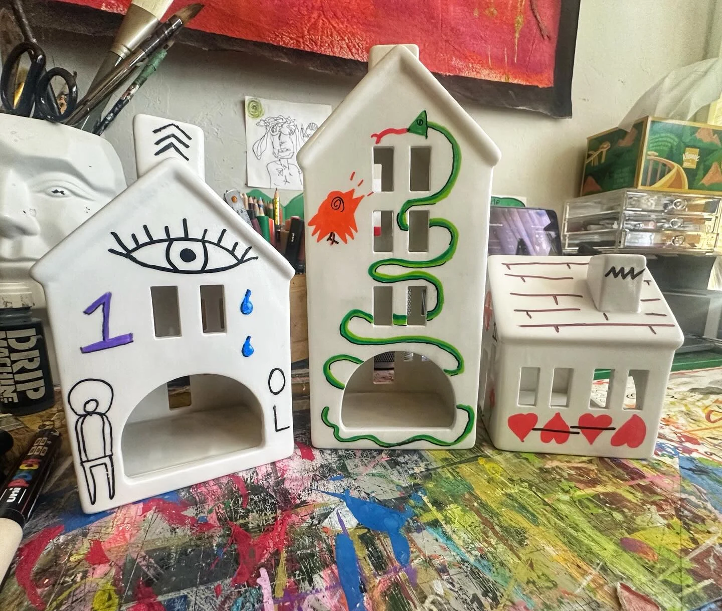 Temples of the Absurd. Three small white house-shaped objects decorated with an eye, a green winding symbol, red hearts, and other painted marks.