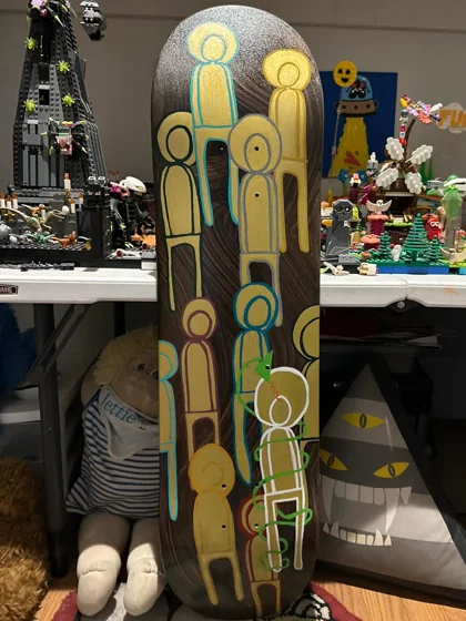 Shared Lines. A skateboard deck covered with overlapping yellow and green human figures on a dark background.